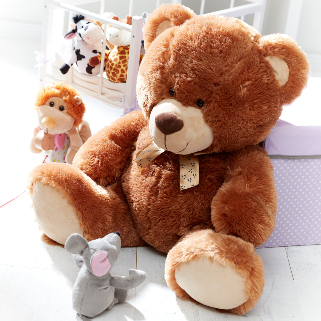 Ein brauner Kuschelbär liegt angenäht an einem Kinderbett. Vor ihm sitzt eine kleine Maus und schaut ihn an. links vom Bären sitzt ein kleiner Löwe.