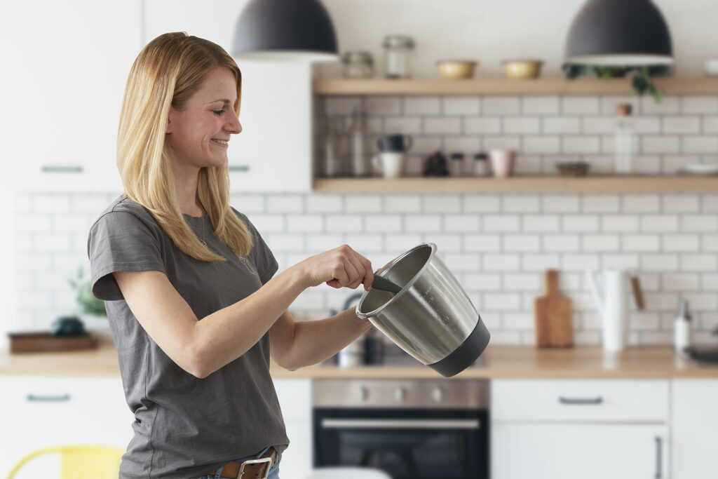 Eine Frau holt mit einem Schaber die letzten Reste aus der Küchenmaschine heraus. Sie steht vor einer modernen lichtdurchflutet Küche.