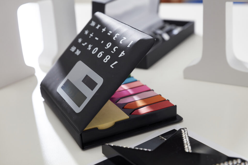 zu sehen, ist ein postit Halter, der sich in einem Taschenrechner verbirgt. Die Post its sind alle in verschiedenen Formen und Farben darunter versteckt. Der Rechner liegt auf einem Schreibtisch.