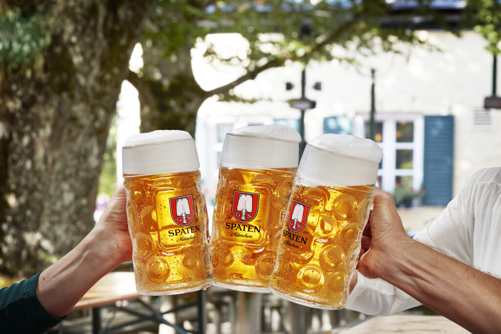 Drei Personen stoßen mit einem Maßkrug in einem Biergarten gemeinsam an. Bei der Bier, einem bayerischen Bier.