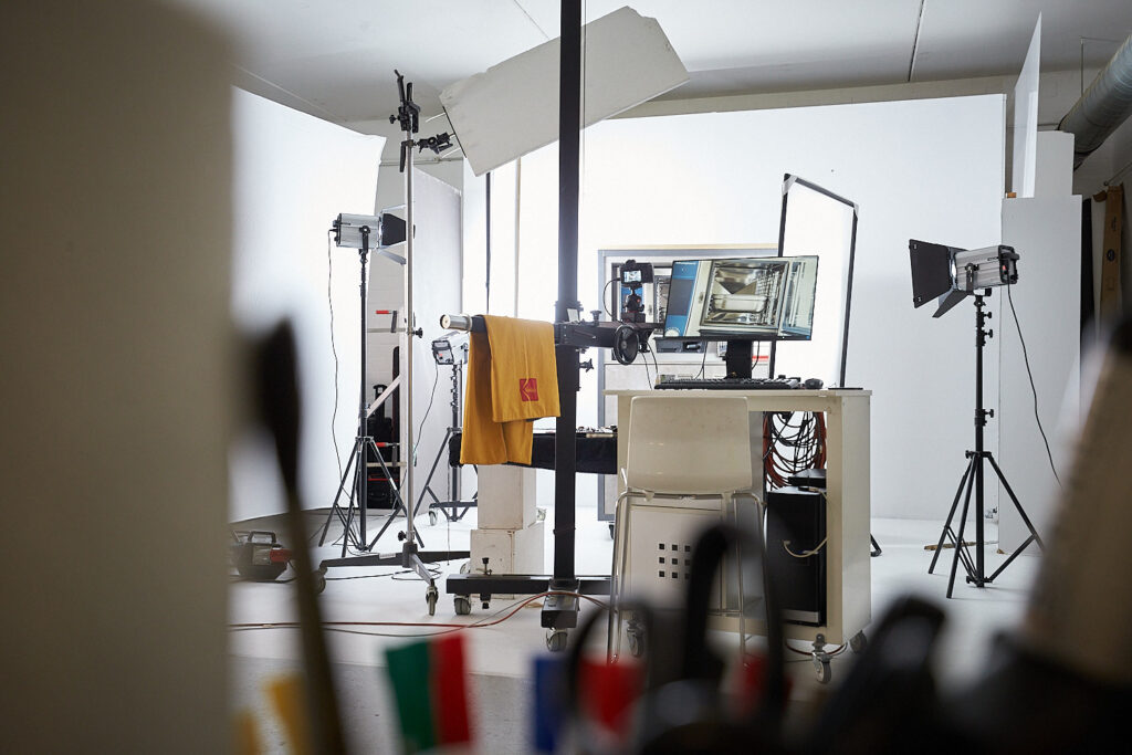 Hier wird als Detailaufnahme der Innenraum des Studios Thorsten Springer gezeigt. Viele Film Lampen erhellen das Studio. In der Mitte des Studios steht ein Tisch mit dem Rechner und zwei Bildschirmen, auf denen man das fertige Foto sehen kann.