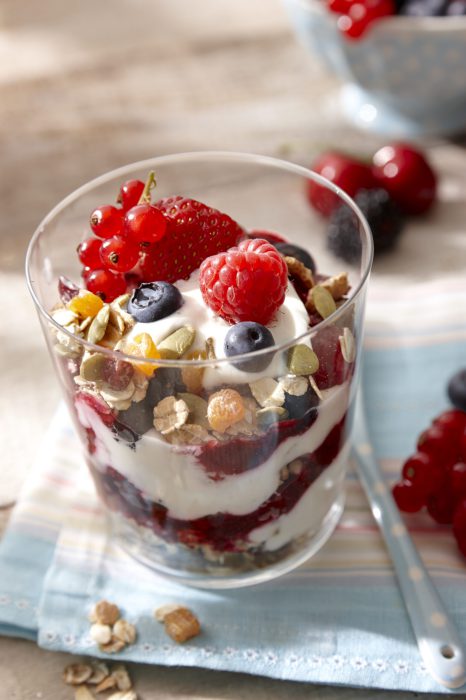 Frühstück im Glas: Joghurt Creme paart sich im Glas mit Haferflocken,Nüssen und Früchten. Das Glas steht im sonnigen Ambiente auf einer Holzplatte.